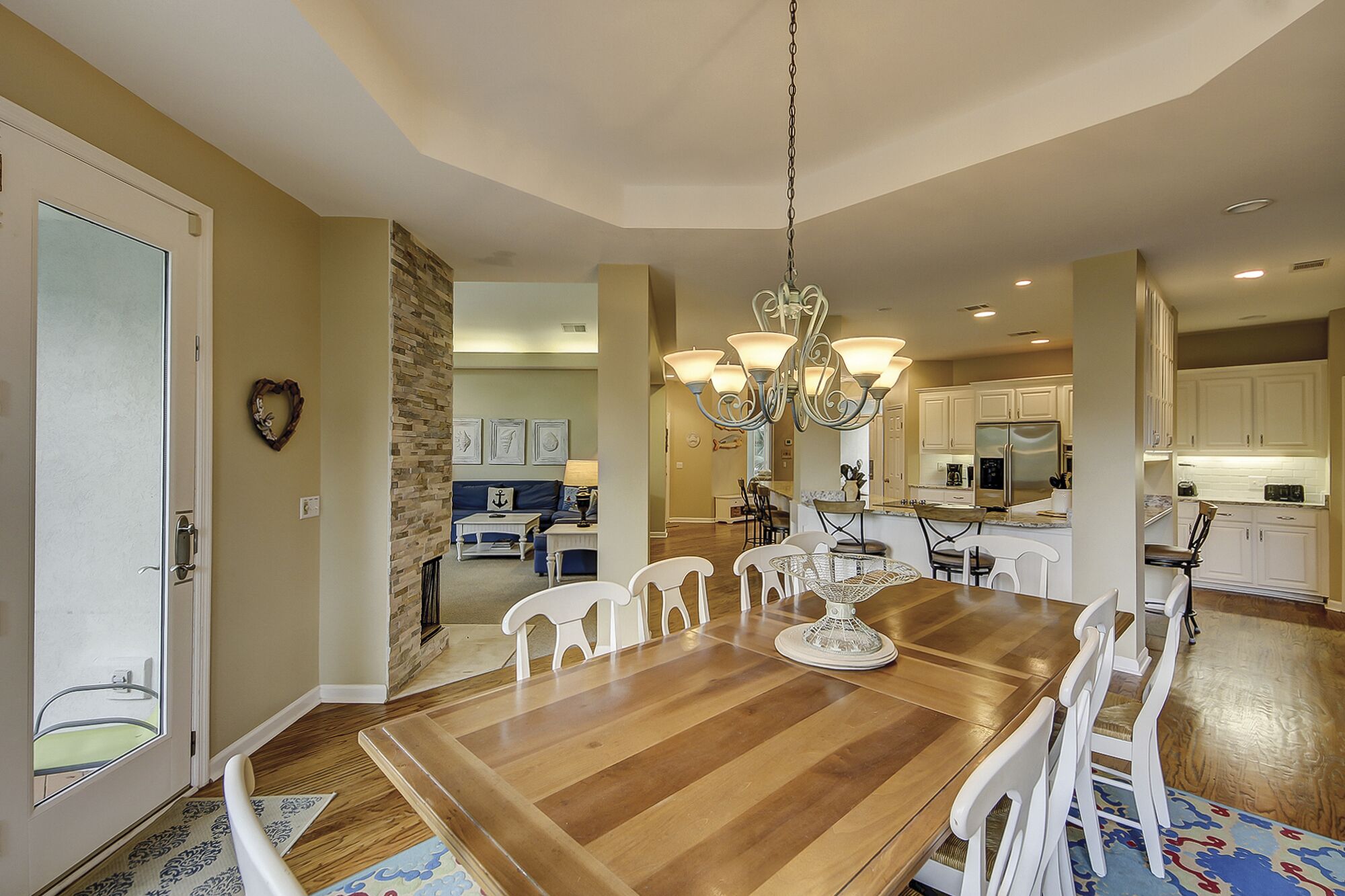 A spacious dining area with a wooden table and white chairs, a chandelier, and a kitchen in the background.