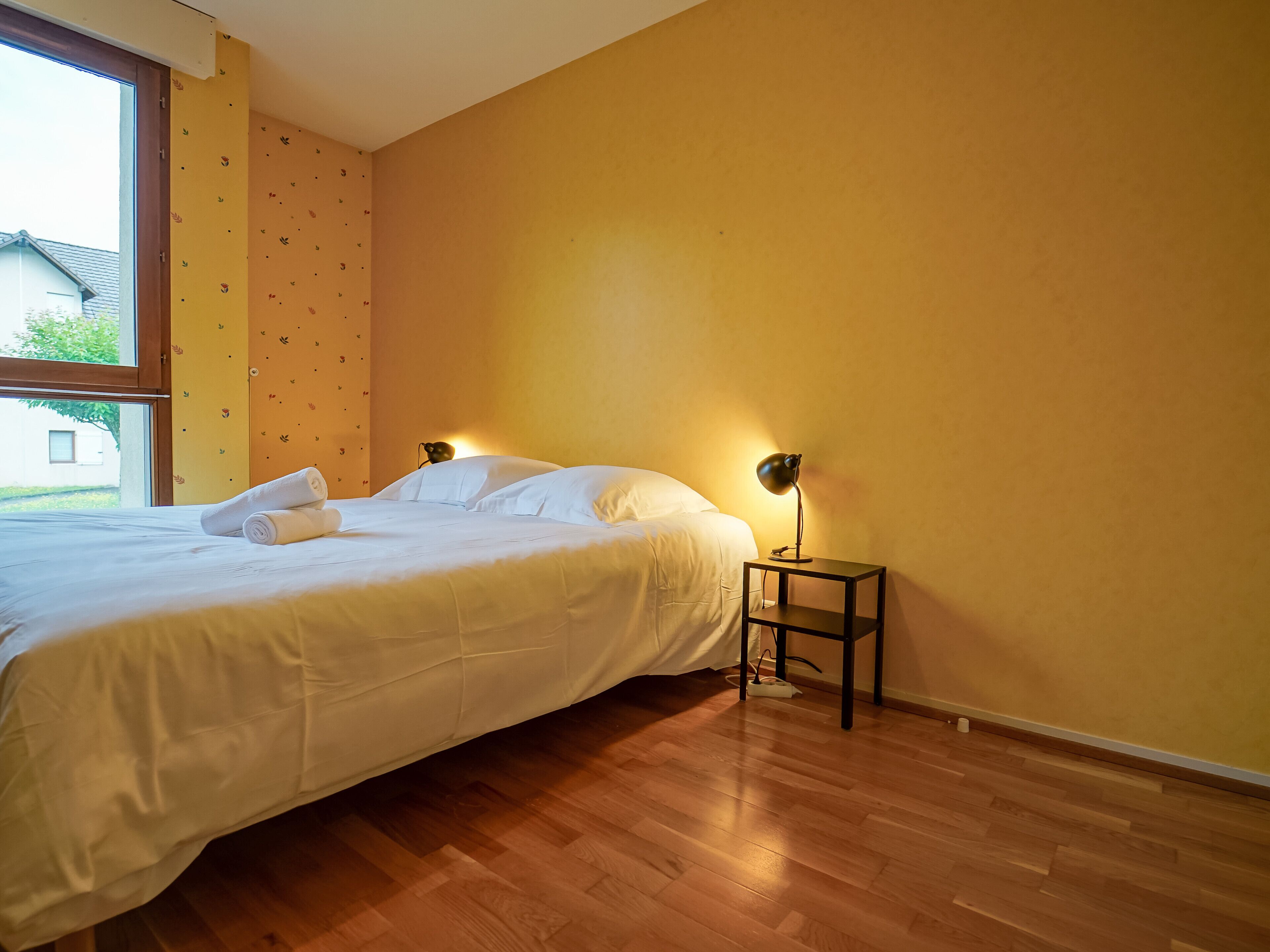 Un dormitorio con una cama, una mesita de noche y una lámpara, y un ventanal.