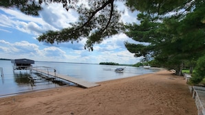 A sandy beach with a dock extending into calm water, surrounded by trees and a clear sky.