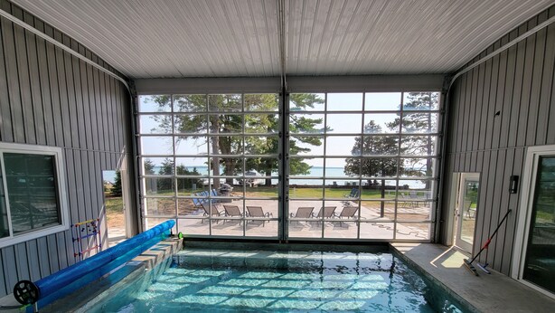 An indoor pool with clear views of the outdoors, including a patio area with chairs and a view of the water.