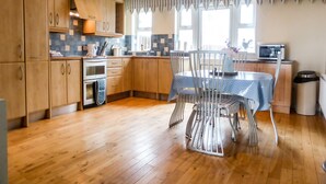 Een keuken met houten kastjes, een eettafel met stoelen, een magnetron en een afvalbak.