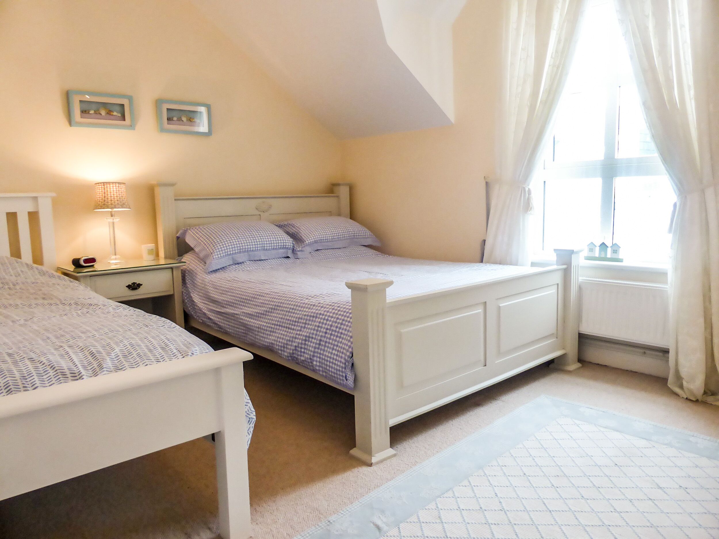A bedroom with two single beds, a bedside table with a lamp, a small shelf with decorative items, and a window with curtains.