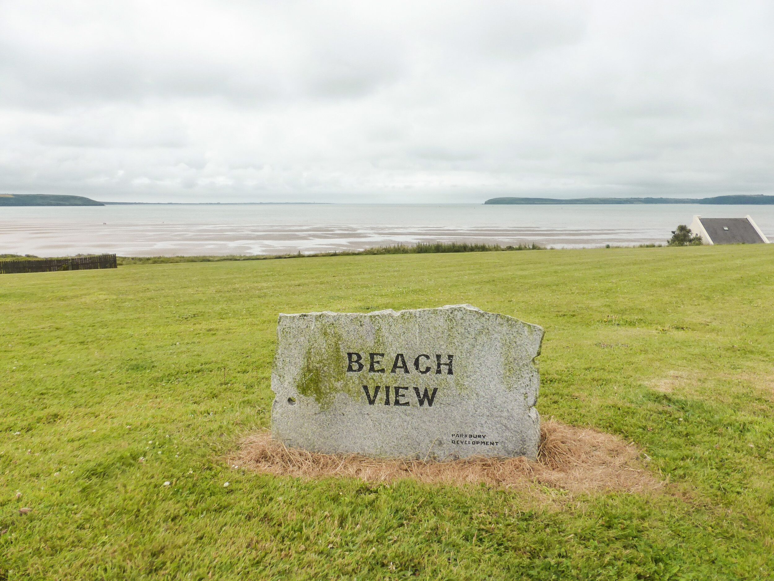A stone sign with "BEACH VIEW" inscribed, surrounded by grass with a view of a body of water and distant land.