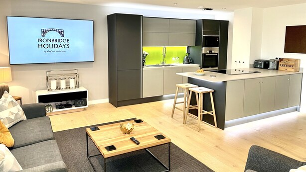 A modern kitchen with grey cabinets, a built-in oven, and a coffee machine. There is a TV mounted on the wall displaying 'Ironbridge Holidays by eley's of ironbridge'. A wooden coffee table with a remote control and a smartphone is in the living area. - Apartment Twelve (Shrewsbury)