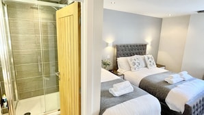 A modern bedroom with a glass-enclosed shower, two beds with grey and white bedding, and a tufted headboard. - Apartment Twelve (Shrewsbury)