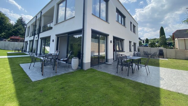 A modern two-story house with large windows, a patio with outdoor furniture, and a well-maintained lawn. - Apartments-Saaldorf WG1 with terrace and view of the reservoir (Bad Lobenstein)