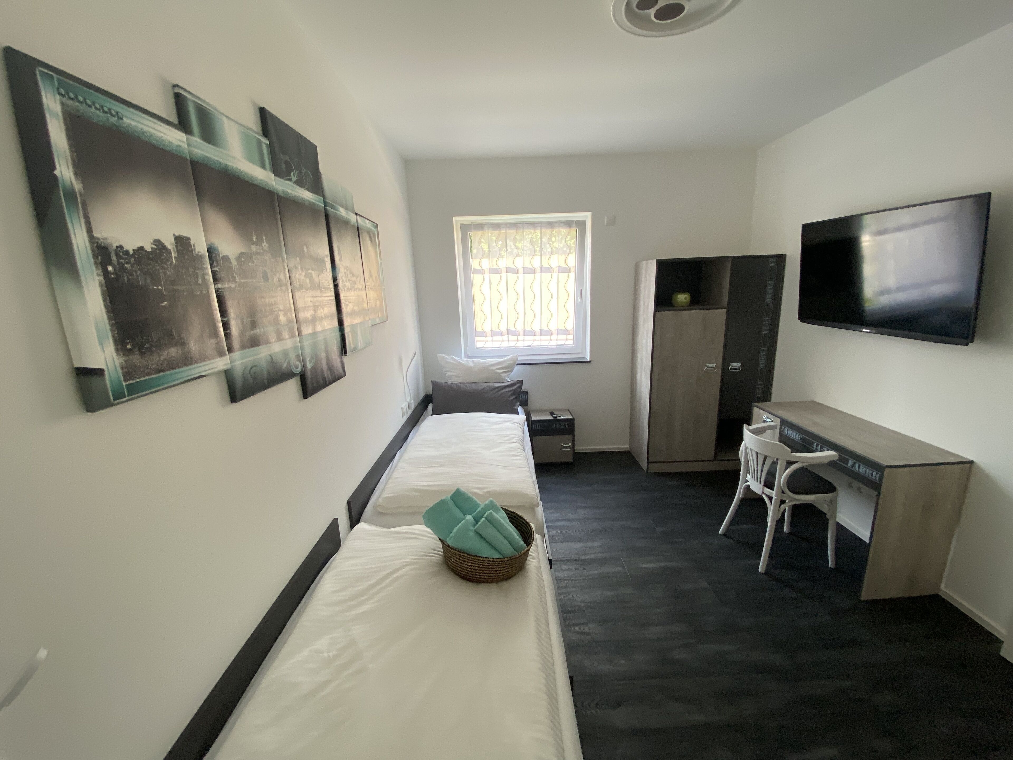 Une chambre d’hôtel de taille réduite, comprenant un lit, un bureau, une chaise, une armoire, un téléviseur et des œuvres d’art accrochées aux murs.