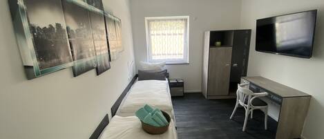 Une chambre d’hôtel de taille réduite, comprenant un lit, un bureau, une chaise, une armoire, un téléviseur et des œuvres d’art accrochées aux murs.