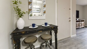 Um corredor com um console de madeira escura, dois banquinhos, um vaso branco com plantas verdes, um espelho com moldura geométrica e uma porta que dá para outro cômodo.