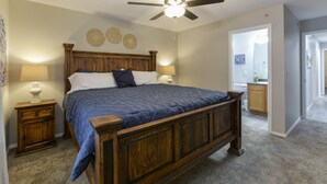 Quarto com cama de madeira, duas mesas de cabeceira, ventilador de teto e banheiro visível por uma porta aberta.