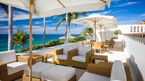 A sunlit outdoor seating area with wicker furniture and white cushions, overlooking a coastal view with palm trees and the ocean. - Luxurious 7 BR Beachfront Casa Oliver w/ WiFi, Pool, Home Theatre, Gym + More! (San Jose del Cabo)