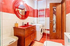 A bathroom with a red and white tiled wall, a wooden vanity with a red sink, a white washing machine, and a wooden door with a towel rack. - Athens Strawberry Hideaway - City Cozy Gem (Athina)
