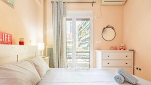 Et soveværelse med seng, natbord, kommode, spejl og udsigt til en balkon med bygninger.