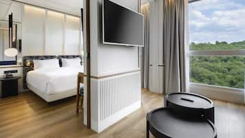 Habitación de hotel moderna con una cama grande, un televisor de pantalla plana, una mesa redonda y vistas al bosque a través de un ventanal amplio.
