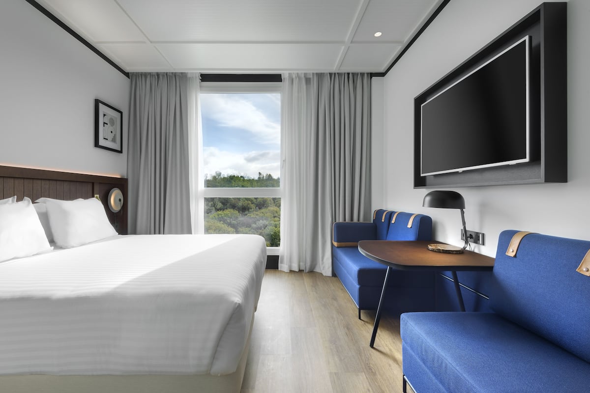 Une chambre d’hôtel comprenant un lit, un coin salon avec une table, un téléviseur fixé au mur et une fenêtre avec des rideaux.