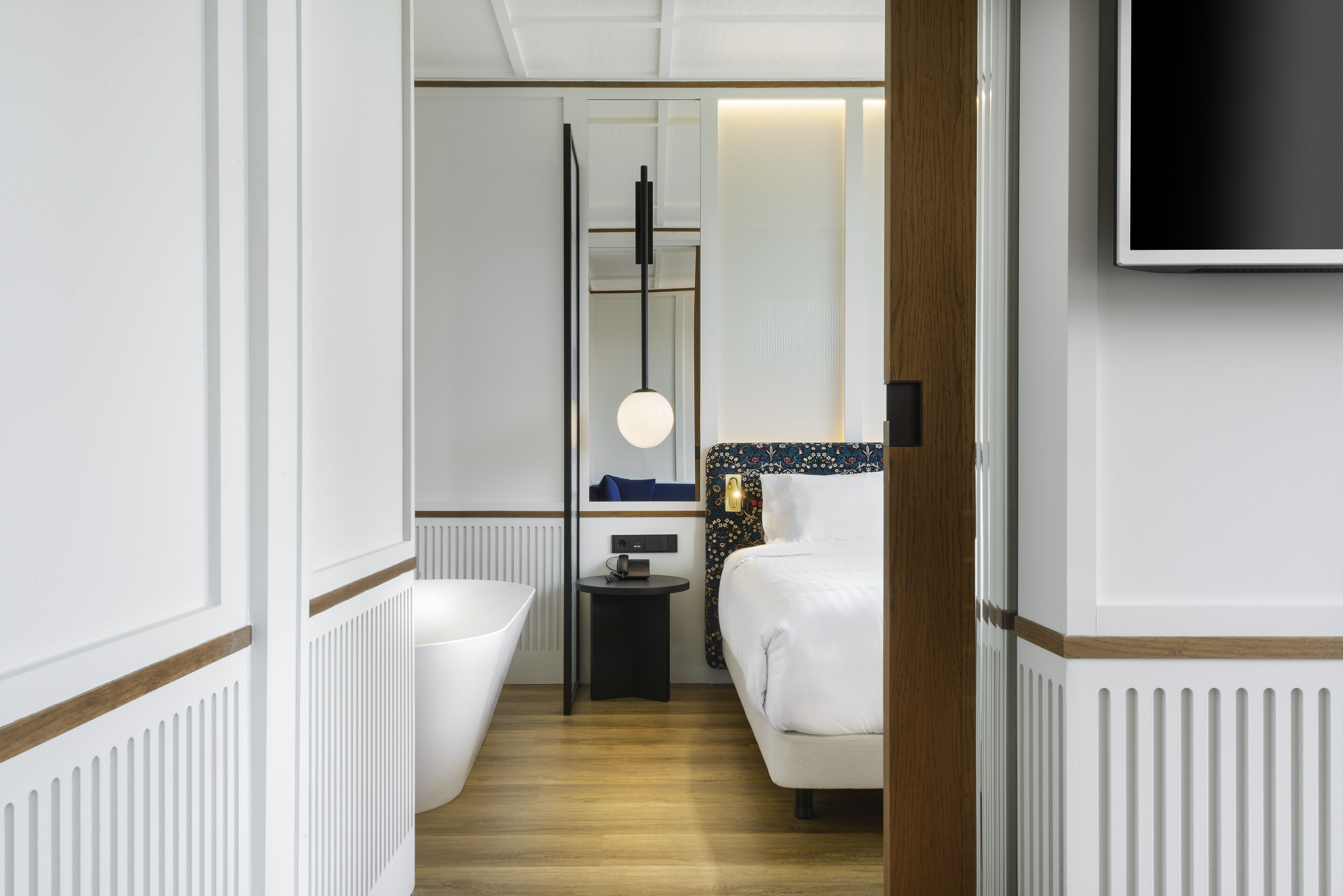 Ein modernes Hotelzimmer mit einer großen Badewanne, einem kleinen Nachttisch, einem flachbildschirmfernseher an der Wand und Holzboden.
