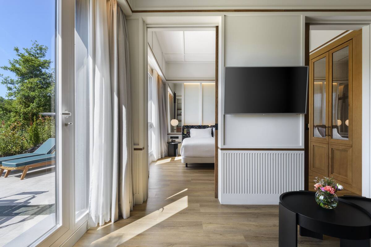 Une chambre d’hôtel moderne dotée d’une grande fenêtre, d’un téléviseur à écran plat, d’un lit et d’une petite table sur laquelle est posé un vase de fleurs.