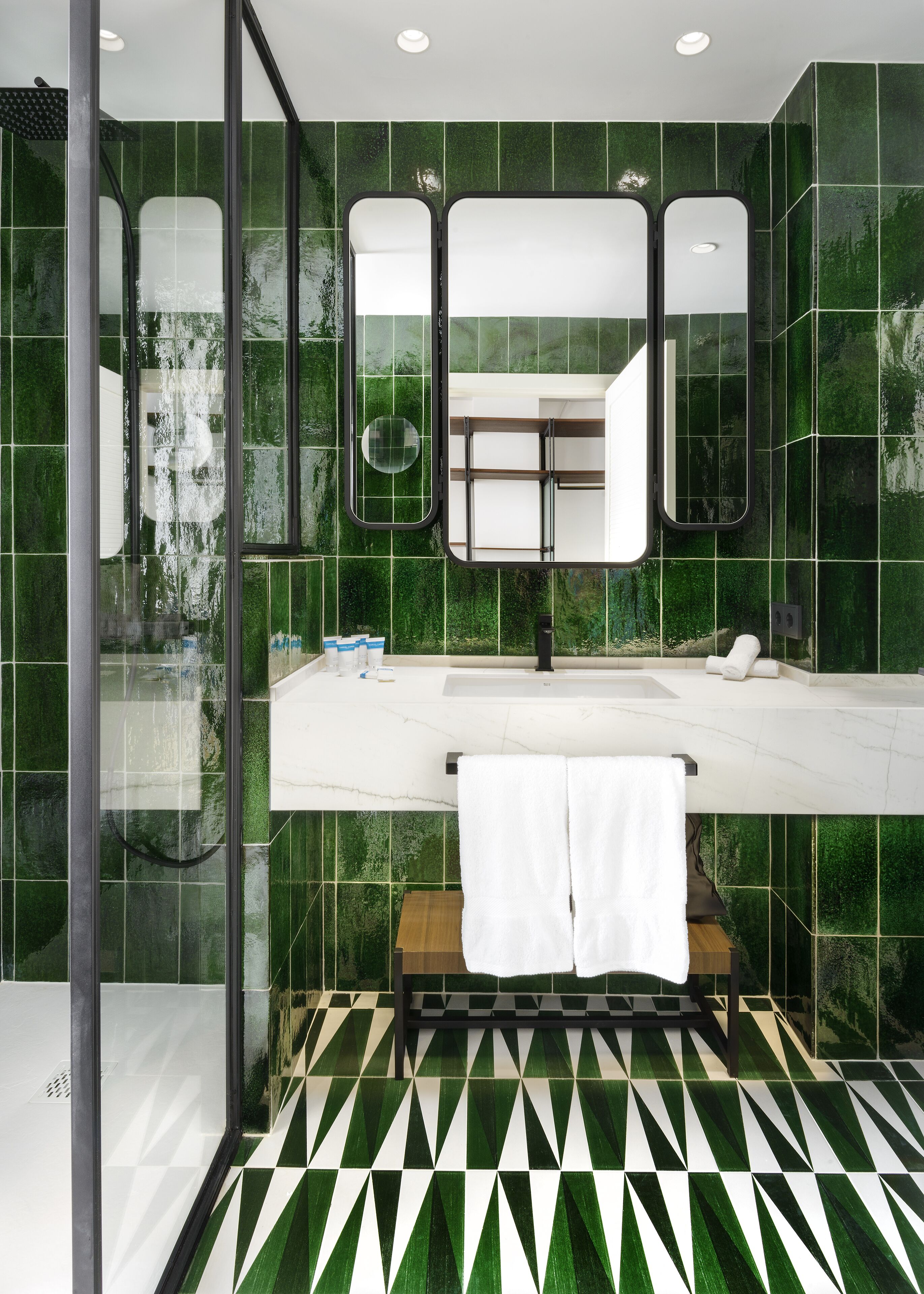 A modern bathroom with green tiles, a large mirror, a white countertop, and a geometric patterned floor.
