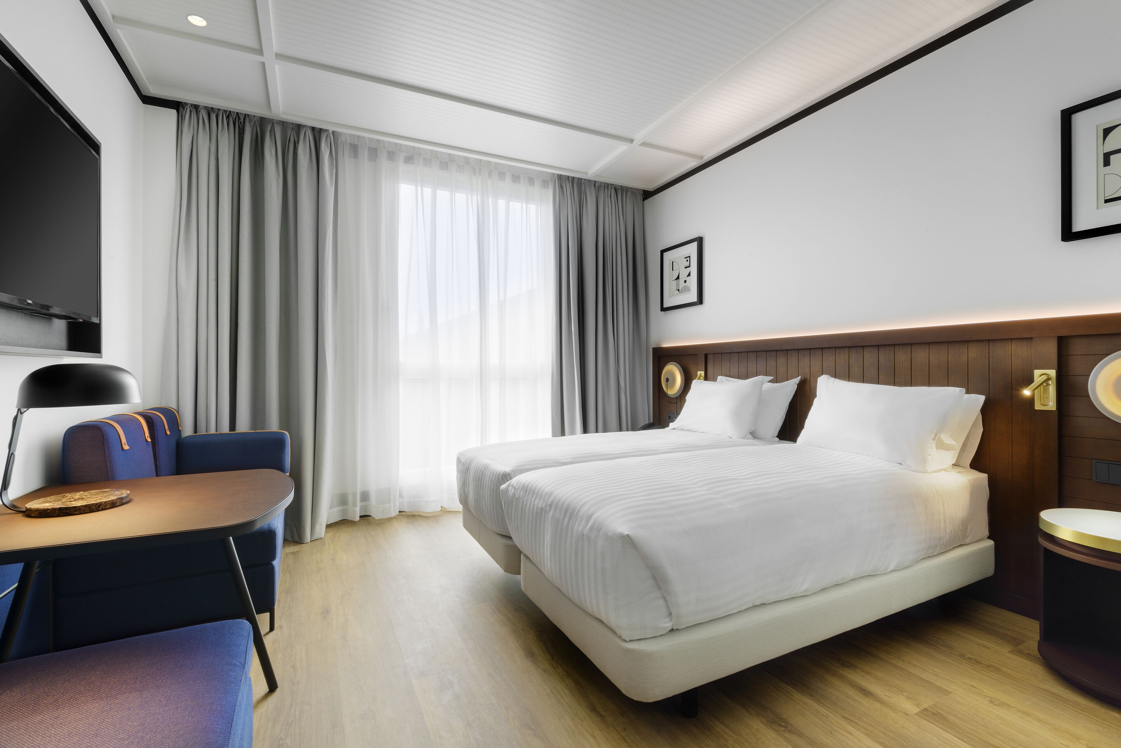 Quarto de hotel moderno com uma cama grande, uma escrivaninha com cadeira, televisão de tela plana e uma janela ampla com cortinas.