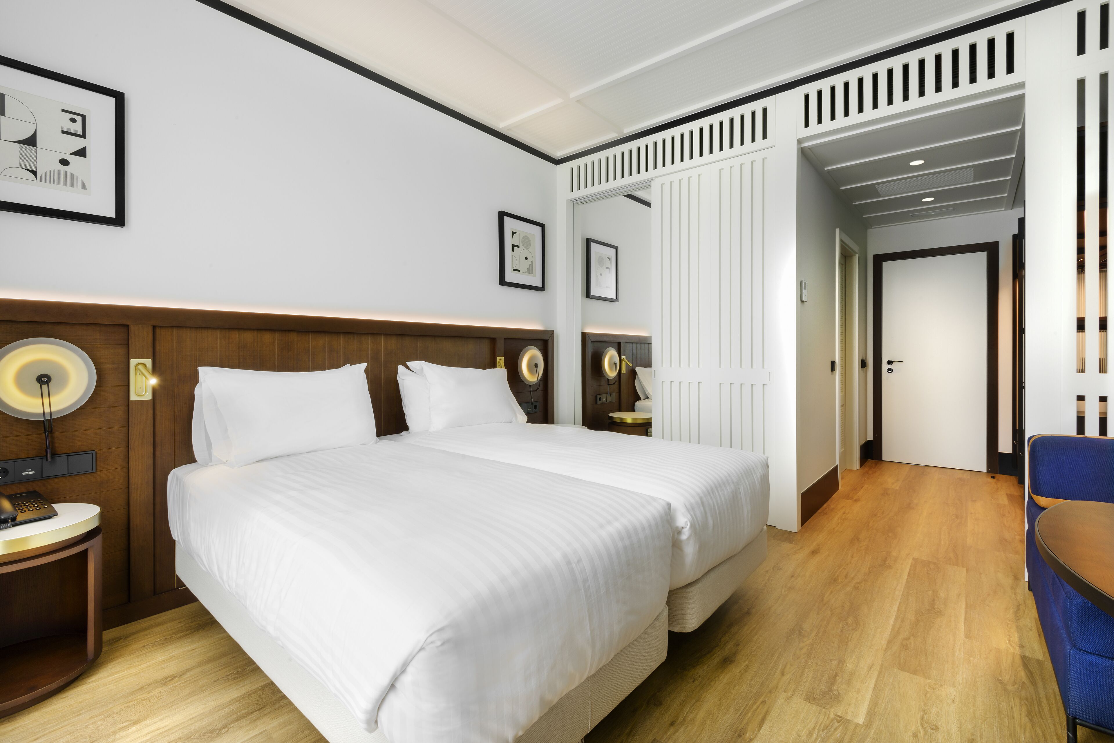 A hotel room with a large bed, bedside tables with lamps, a wooden headboard, and a door leading to another room.