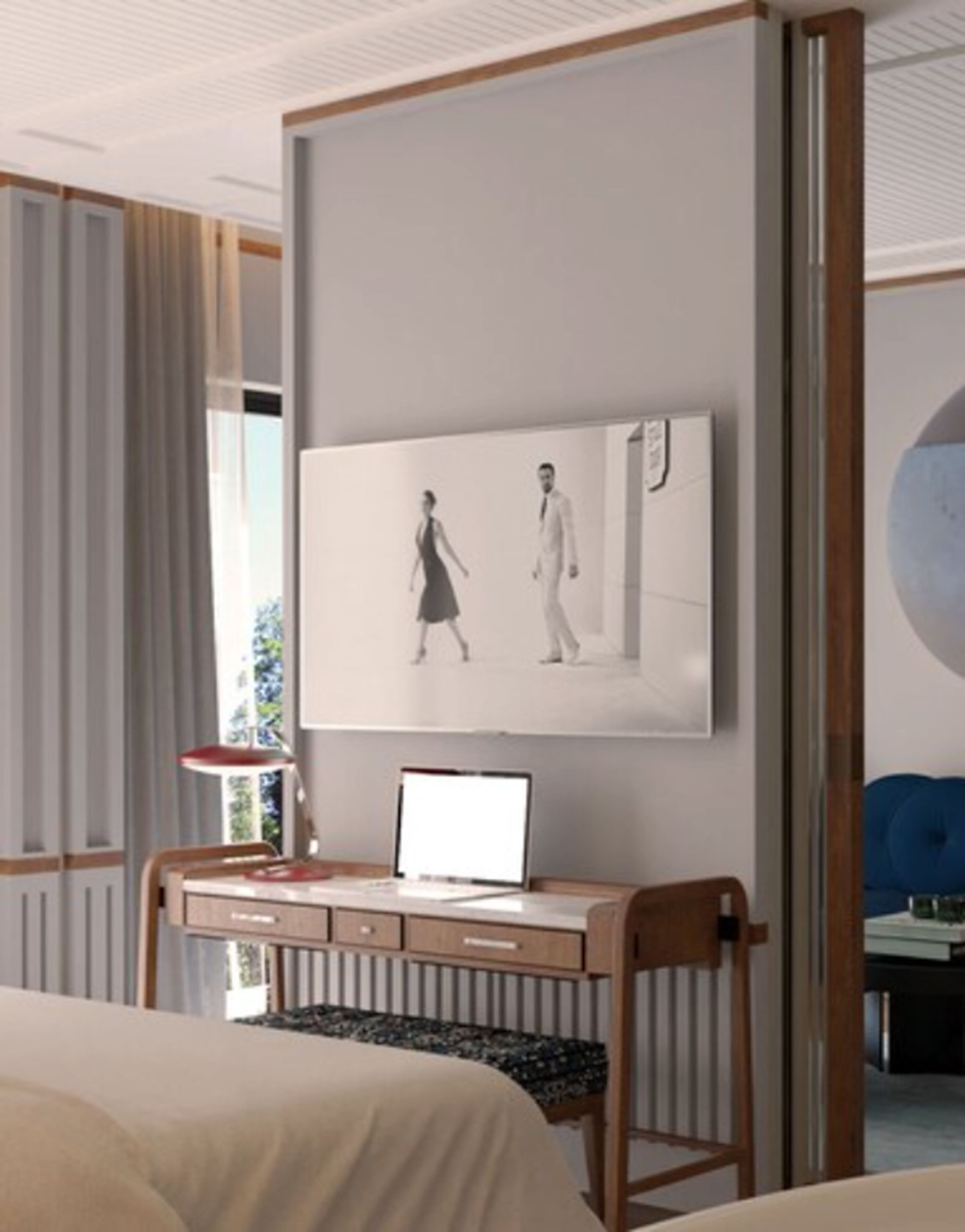 A modern hotel room with a bed, a desk with a laptop, a chair, and a large wall-mounted TV displaying a couple walking.
