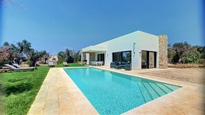 Una casa moderna con piscina, mobiliario exterior y un muro de piedra.
