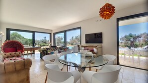 A modern living room with a glass dining table surrounded by white chairs, a large sofa, and a unique hanging light fixture.