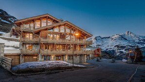 A multi-story wooden chalet with balconies, set against a snowy mountain backdrop.