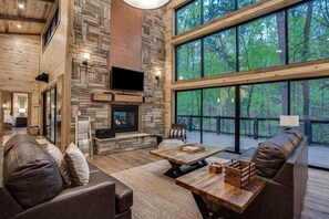 A spacious living room with a stone fireplace, leather sofas, and large windows overlooking a forest.