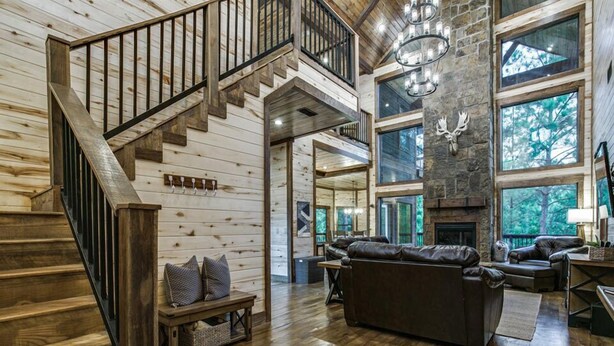 A spacious living area with a wooden staircase, leather furniture, and large windows offering a view of the outdoors. - "Rustic Mountain Lodge" Bunk Room, Outdoor Entertaining, Pets (Broken Bow)