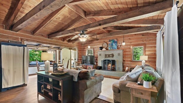 A cozy cabin interior with a wooden ceiling, a fireplace, a sofa, a dining area, and a view of the outdoors.