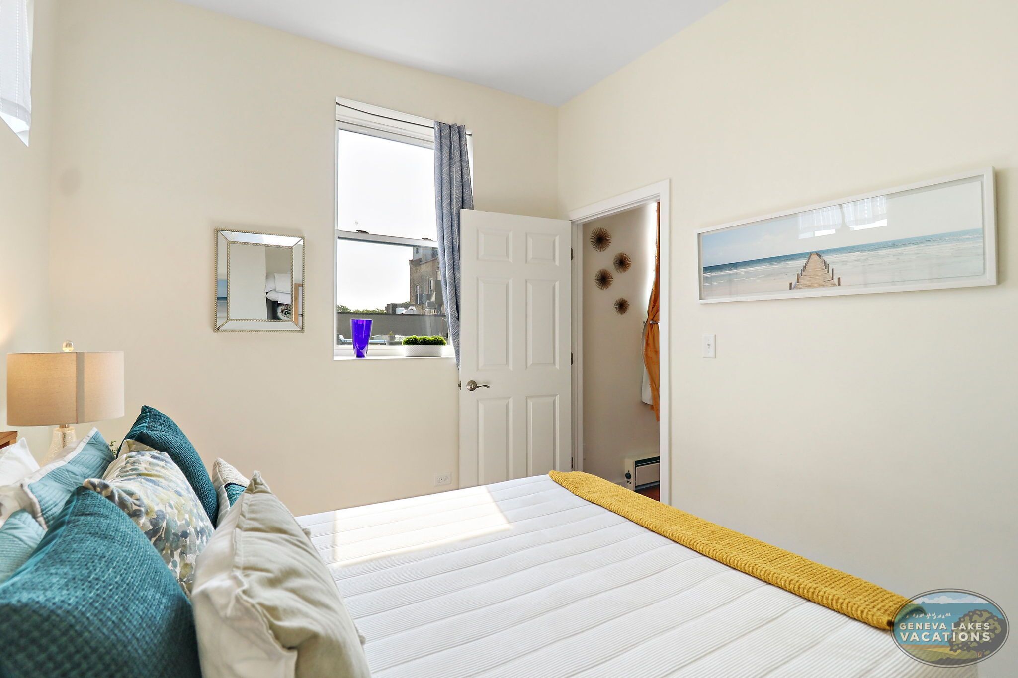 Un dormitorio con cama, ventana, puerta, espejo y un cuadro en la pared.
