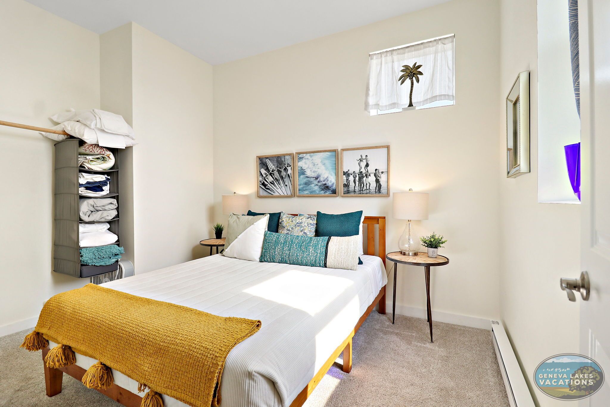 A neatly made bed with a white comforter and a mustard-colored throw blanket, flanked by two bedside tables with lamps and a small plant. A wall-mounted shelf holds folded towels and a white robe. Three framed artworks adorn the wall above the bed, and a window with a palm tree curtain is visible.