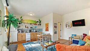 Un salon moderne avec un canapé, une table basse et un coin repas. On aperçoit une cuisine à l’arrière-plan, avec des meubles en bois et une décoration sur le thème du surf.