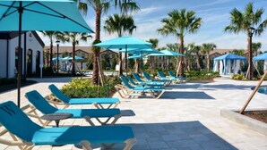 Zona junto a la piscina con sillones de descanso y sombrillas, rodeada de palmeras y edificios.