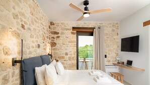 A bedroom with a stone wall, a bed with white linens, a ceiling fan, a TV, and a laptop on a wooden shelf. - Dianthus Residence, Heated Pool & in-pool Whirlpool (Adele)