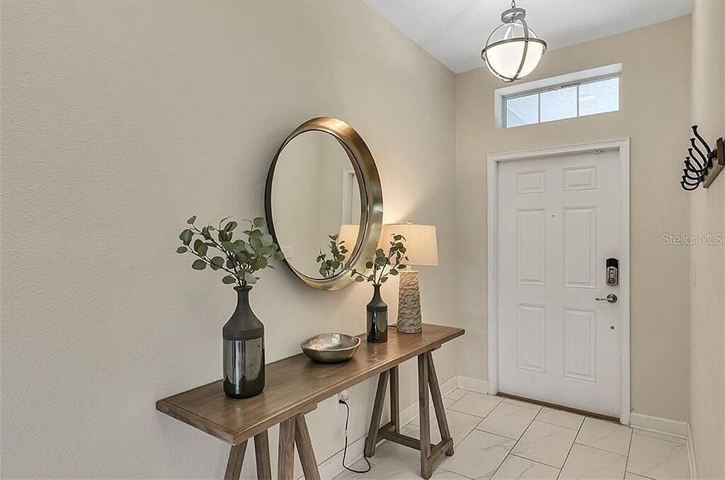 A hallway with a wooden console table, a mirror, a vase with plants, a bowl, a lamp, and a white door.