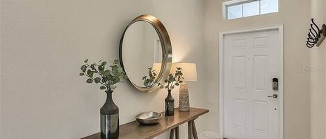 A hallway with a wooden console table, a mirror, a vase with plants, a bowl, a lamp, and a white door.