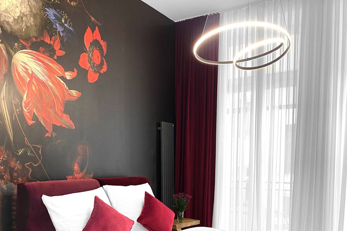 A modern hotel room with a large bed, red and white bedding, a decorative wall mural, a round light fixture, and sheer curtains.