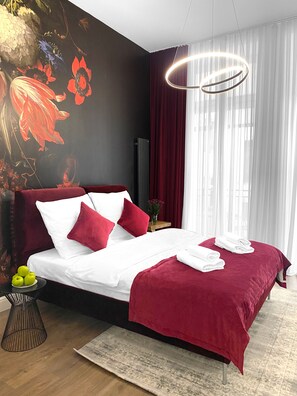 Moderni hotellihuone, jossa on suuri sänky, punavalkoiset vuodevaatteet, koristeellinen seinämaalaus, pyöreä valaisin ja läpikuultavat verhot.