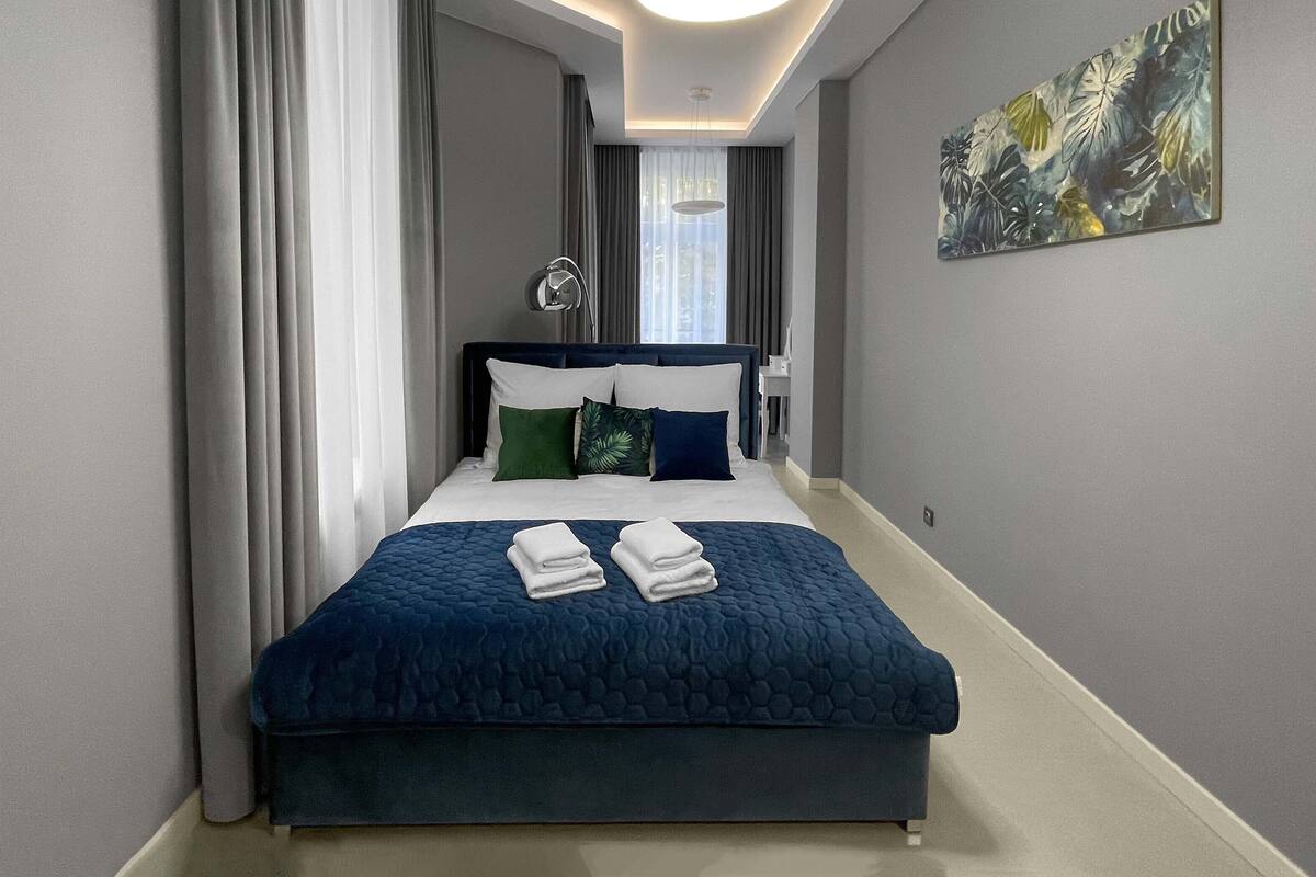 A neatly made bed with a blue quilt and two pillows, a bedside table with a lamp, and a painting on the wall.