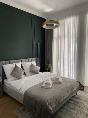 A modern bedroom with a large bed, grey bedding, a bedside table with a vase of flowers, and sheer curtains. - Starowiślna 41-43 by PI Apartments (Kraków)
