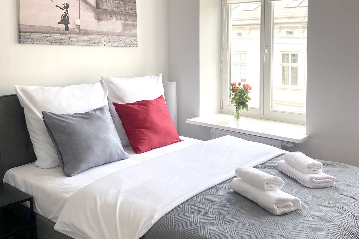 A neatly made bed with white and grey bedding, a red pillow, and a grey pillow. There is a window with blinds, a small table with a vase of flowers, and a painting on the wall.