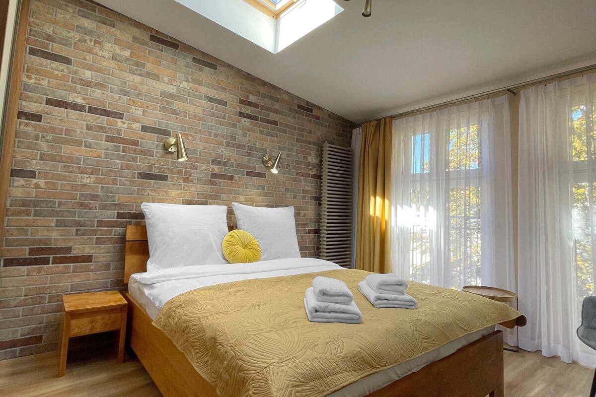 A modern bedroom with a skylight, brick wall, and a bed with white linens and a yellow pillow.