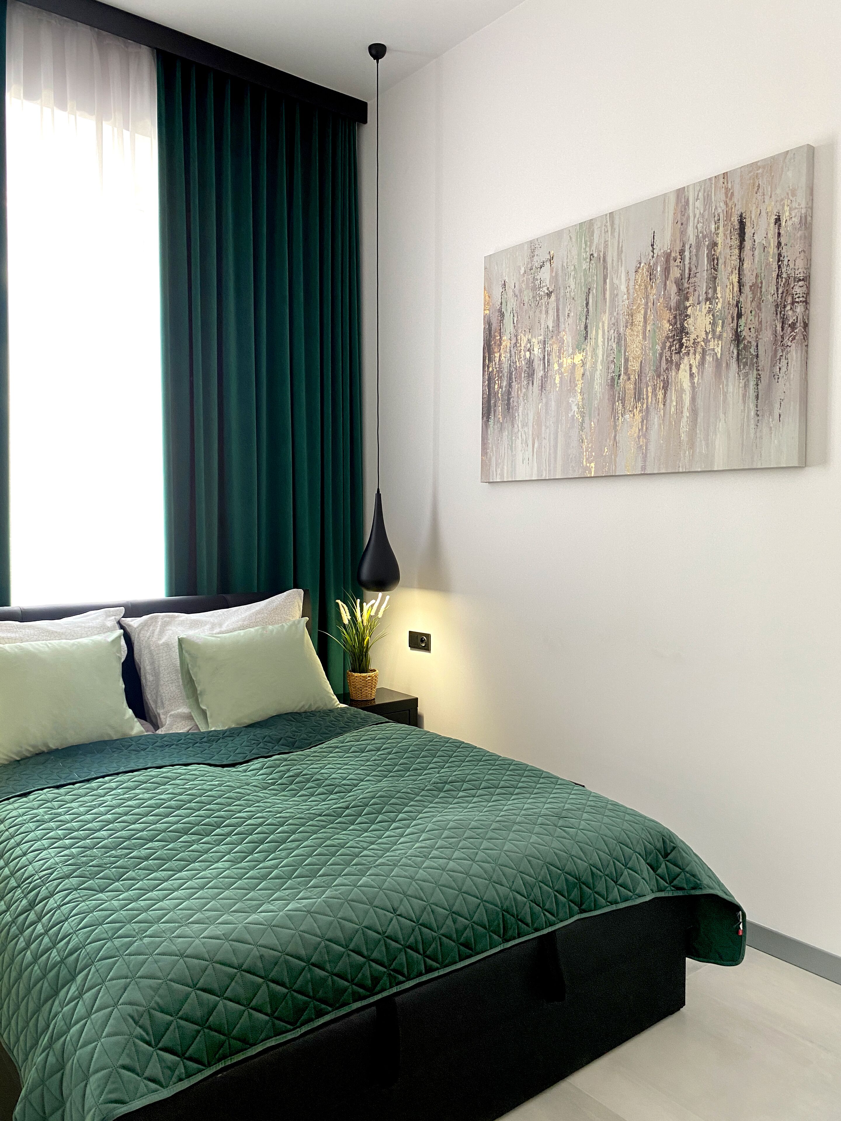 A bedroom with a green quilted bedspread, white pillows, a dark nightstand with a lamp, and a painting on the wall.