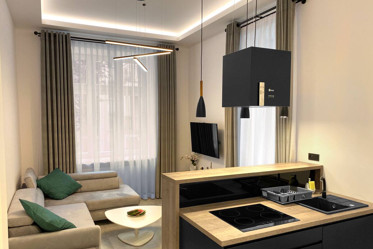 A modern living room with a sofa, a coffee table, a TV, and a kitchen area with a stove and cabinets.
