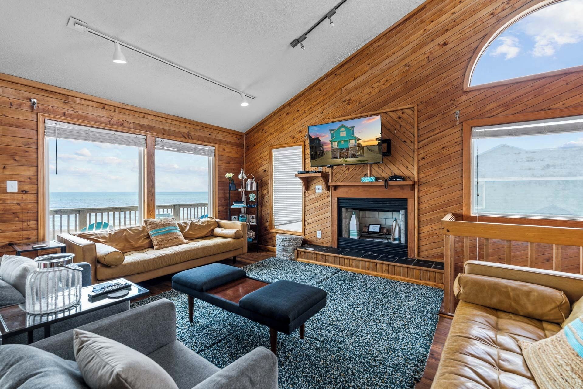A cozy living room with a fireplace, wooden walls, a large window overlooking the sea, and a comfortable seating area.