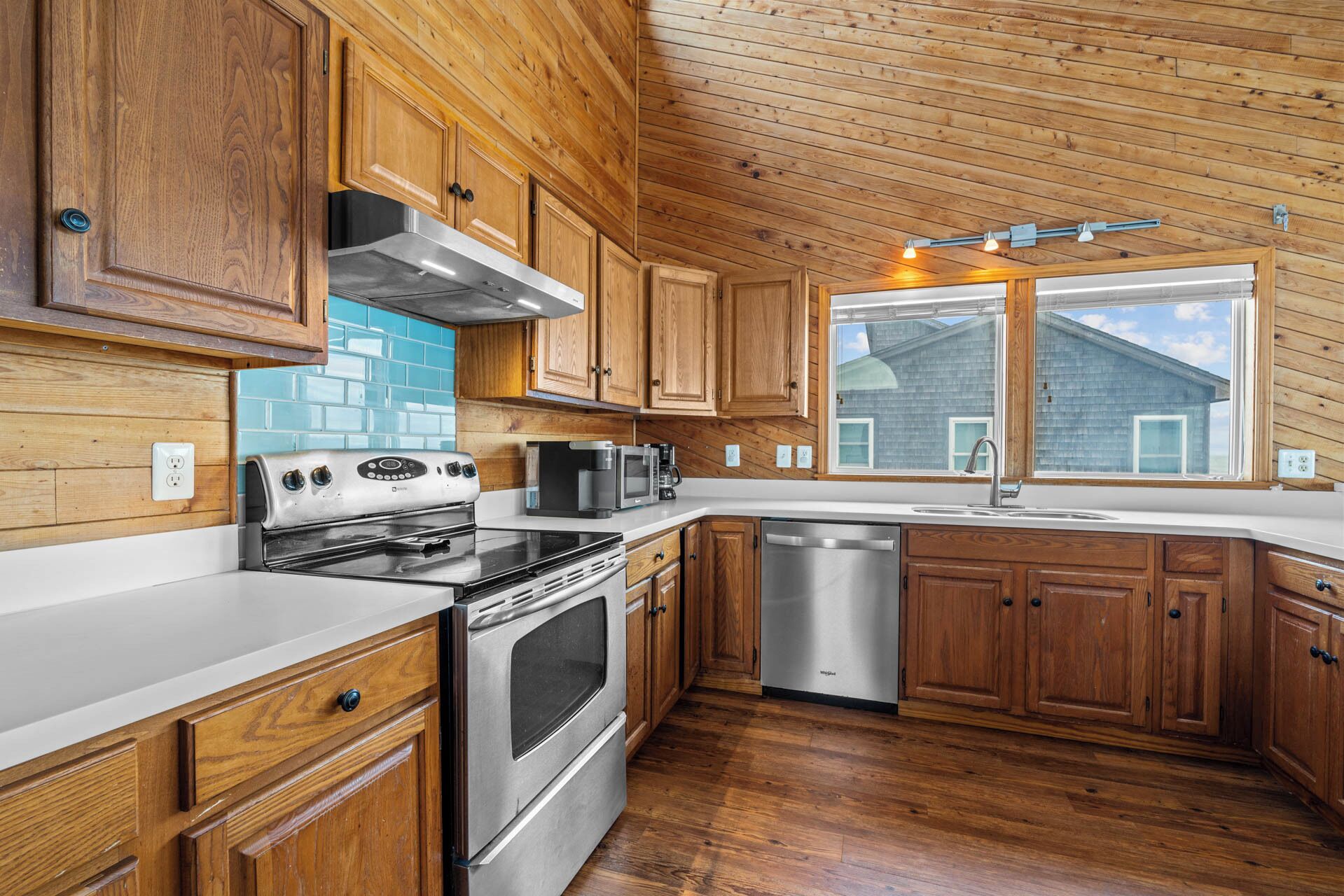 A modern kitchen with wooden cabinets, stainless steel appliances, and a large window offering a view of the house.