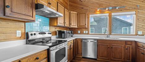 A modern kitchen with wooden cabinets, stainless steel appliances, and a large window offering a view of the house.