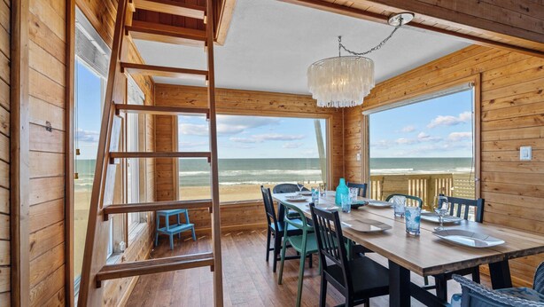 A wooden cabin with a dining area overlooking the beach, featuring a wooden ladder, a chandelier, and a table set for two. - Oceanfront Dog Friendly Home w/ Hot Tub & Grill (Avon)